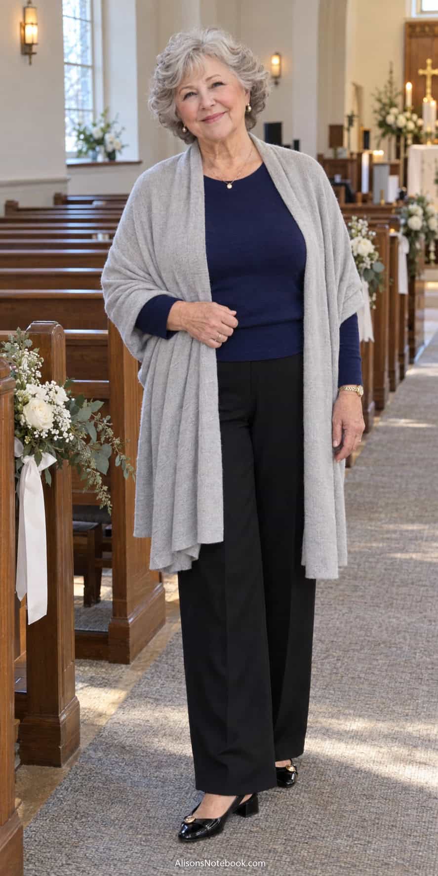 Woman around eighty with short curly silver hair wearing a navy sweater, black wide-leg pants, a light gray shawl, and black low-heeled shoes in a church decorated for a wedding.