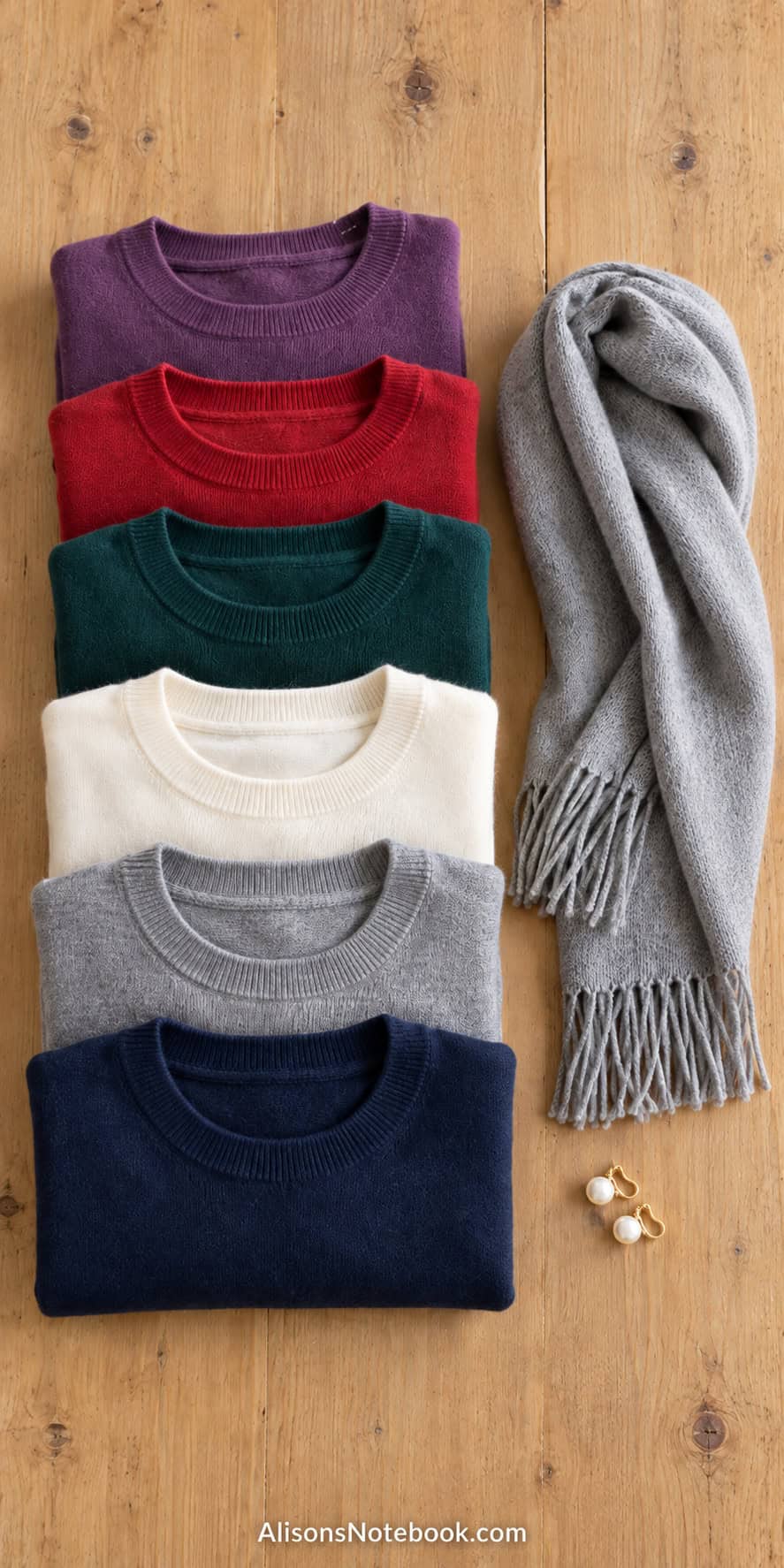 Folded soft sweaters in purple, red, deep green, cream, gray, and navy with a gray scarf and pearl earrings on a wood table.