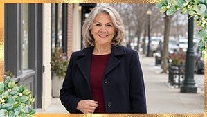 Woman around seventy with silver hair in a navy wool coat over a burgundy knit dress on a winter town sidewalk, framed in gold with green leaves