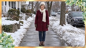 Smiling woman in her 60s in a wine-red wool coat, ivory scarf and cream beanie on a snowy small-town sidewalk