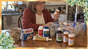 A woman in her 60s in a rust cardigan reads a prescription bottle beside Tylenol, Advil, Centrum and Band-Aids on a table