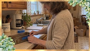 A woman in her 60s in an oatmeal cardigan presses blue painter's tape onto an old loaf pan on a kitchen counter