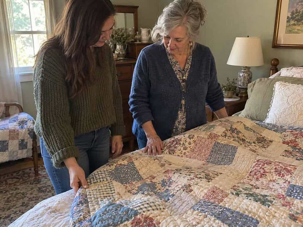A young woman and an older woman looking at a faded handmade quilt on a guest bed, with the young woman touching its edge.